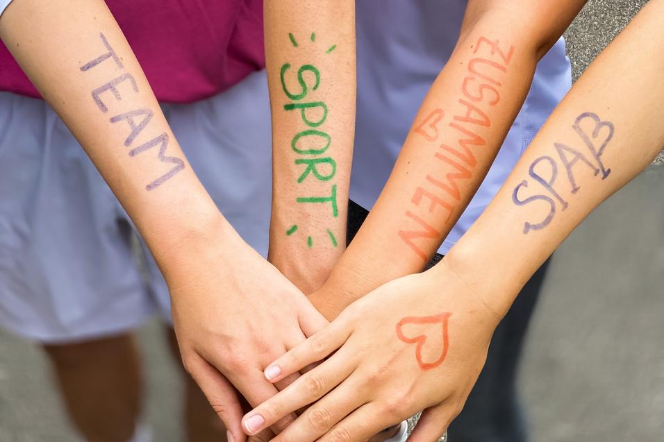 Vier Hände mit bunten Schriftzügen "TEAM", "SPORT", "ZUSAMMEN" und "SPASS" liegen übereinander auf einem grauen Untergrund.