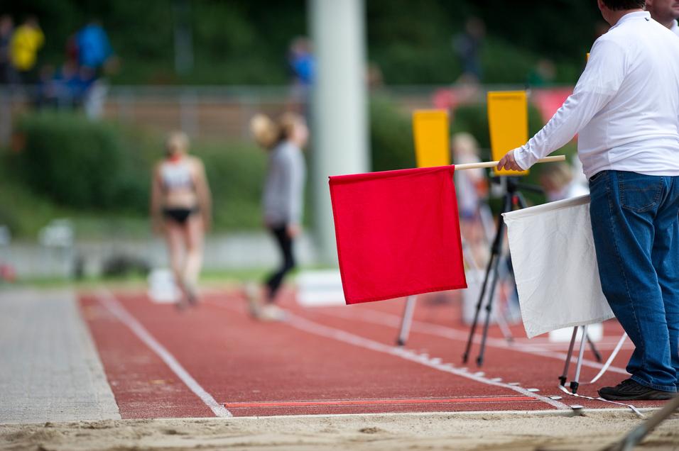 Ein Schiedsrichter hält eine rote Flagge neben einer Laufbahn, während Athleten im Hintergrund vorbeigehen.