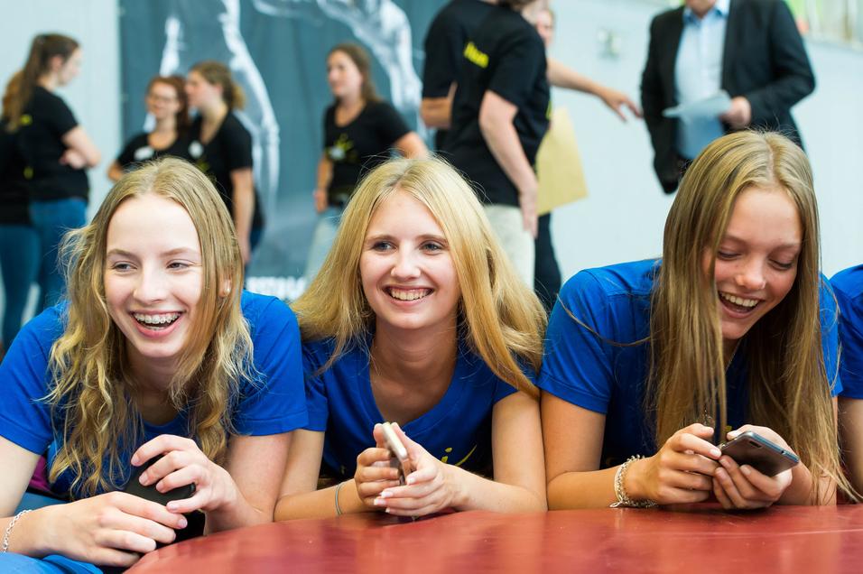 Drei lachende Jugendliche in blauen T-Shirts liegen auf einem Tisch und schauen auf ihre Handys. Hintergrund mit weiteren Personen.
