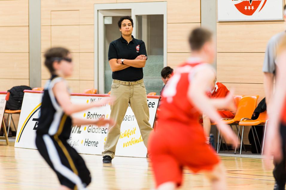 Trainer steht am Spielfeldrand und beobachtet ein Basketballspiel mit spielenden Kindern.